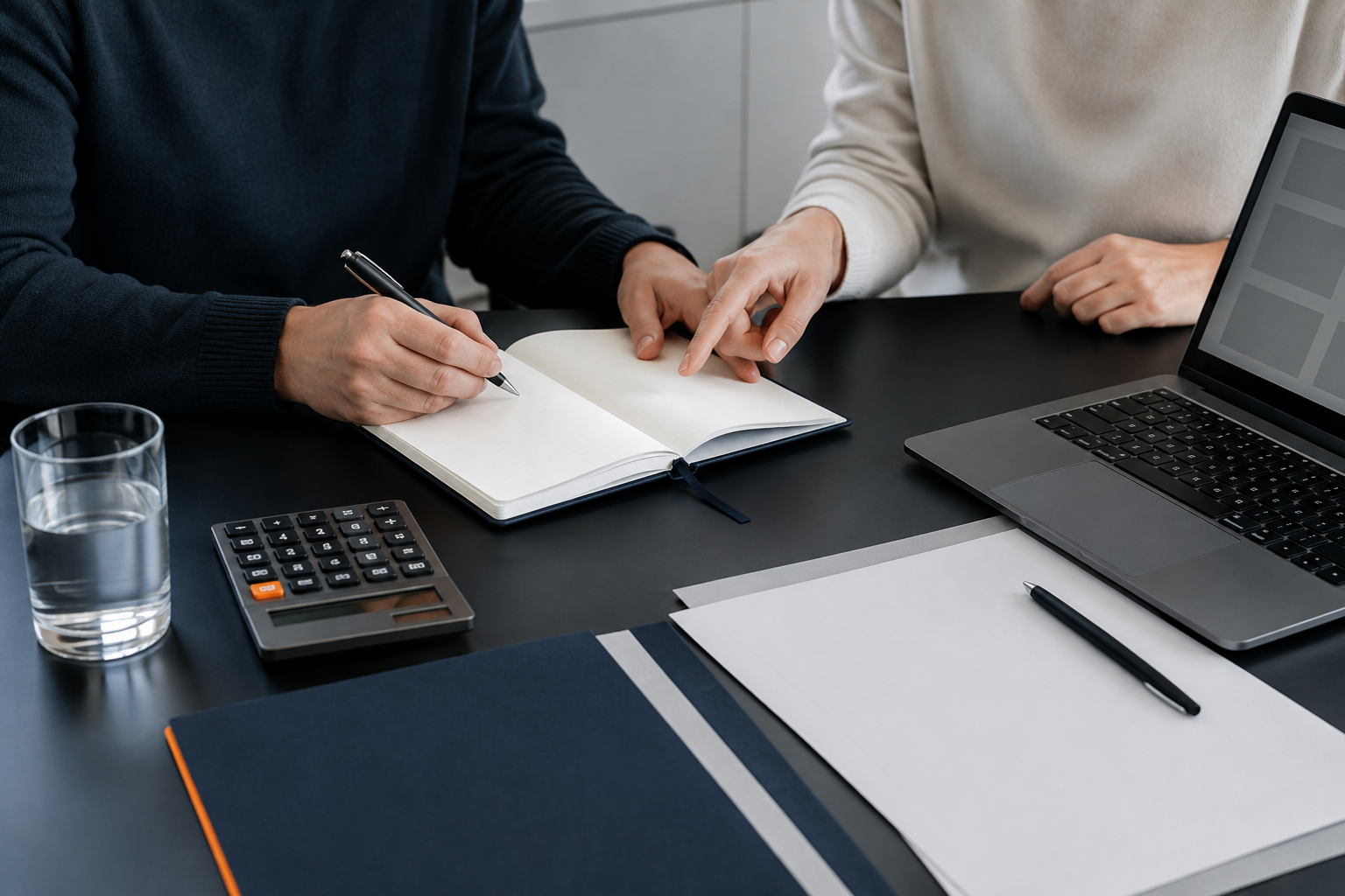 Un carnet et une calculatrice sur une table de travail, avec deux personnes préparant un dossier.